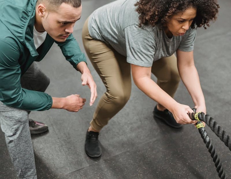 From above of concentrated young male trainer instructing determined obese African American woman doing exercise on battle ropes during functional workout in gym with